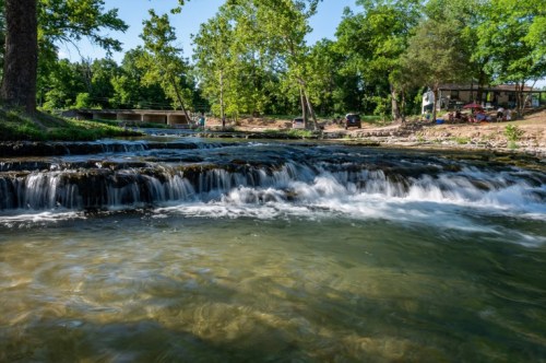 A serene river with gentle waterfalls, surrounded by lush greenery and a shaded area with people in the background.