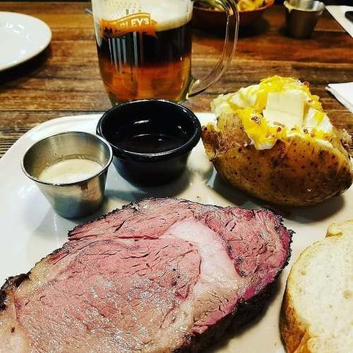 A plate with sliced roast beef, a baked potato topped with cheese, and two dipping sauces, alongside a glass of beer.
