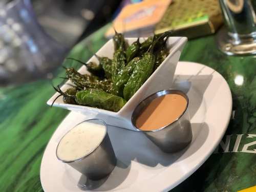 A bowl of grilled green peppers served with two dipping sauces on a white plate.