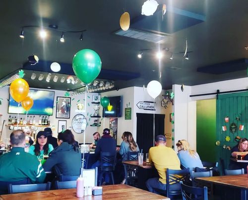 A lively pub interior decorated with green and gold balloons, featuring patrons enjoying drinks and food at wooden tables.