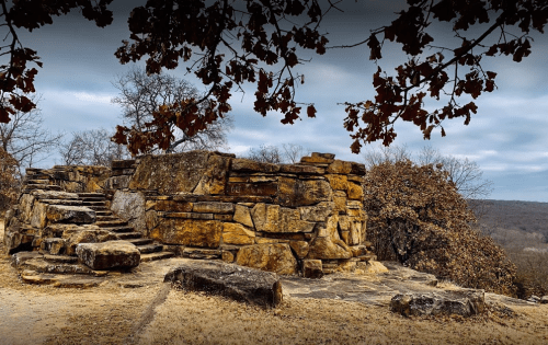Ancient stone ruins surrounded by trees, with a cloudy sky overhead and a rugged landscape in the background.