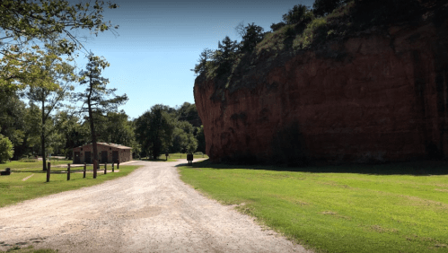 A dirt path leads through a green area with a large rock formation and a small building in the background.