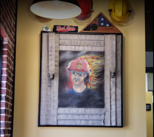 A framed portrait of a firefighter with flames in the background, displayed on a wall with helmets and memorabilia.