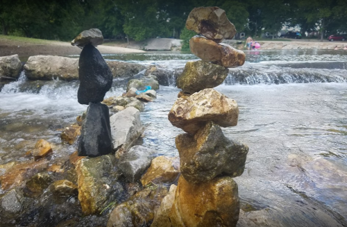 Two rock sculptures balanced on a riverbank, with flowing water and greenery in the background.