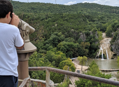 A child looks through a telescope at a scenic view of a waterfall and lush green hills.