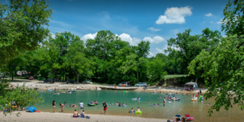 A sunny riverside scene with people swimming and relaxing on the shore, surrounded by trees and a clear blue sky.