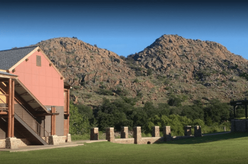 A rustic building with a staircase beside rocky hills and greenery under a clear blue sky.