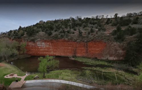 A scenic view of a red rock cliff beside a calm river, surrounded by greenery and trees.