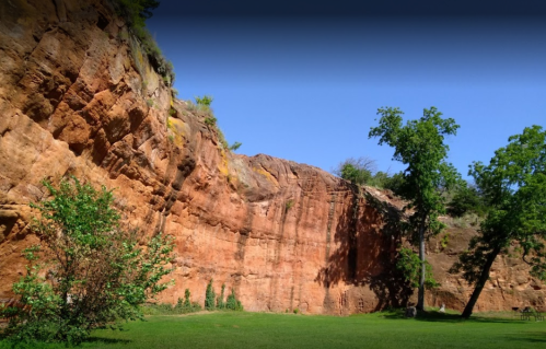 A rocky cliff with reddish hues, surrounded by green grass and trees under a clear blue sky.
