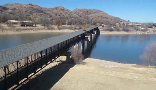 A long wooden bridge spans a calm lake, surrounded by hills and trees under a clear blue sky.
