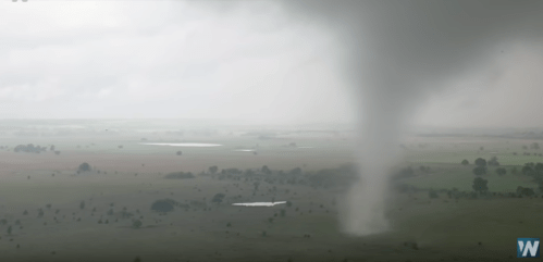 A tornado touches down in a flat landscape, with a cloudy sky and distant fields visible.