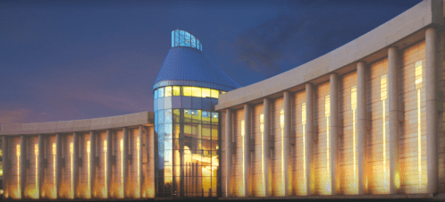 Modern building with a curved facade, large glass windows, and a distinctive pointed roof, illuminated at dusk.