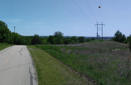 A paved path alongside grassy fields, with power lines and a clear blue sky in the background.