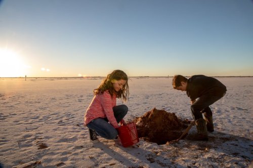 Two children play on a sandy beach at sunset, digging in the sand with a bucket and shovel.