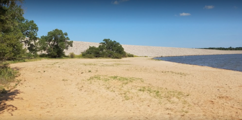 A sandy shoreline with a calm water body and a stone dam in the background under a clear blue sky.