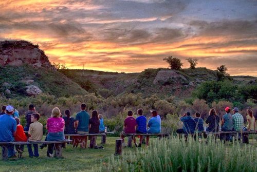 A group of people sitting on benches, watching a colorful sunset over a scenic landscape.