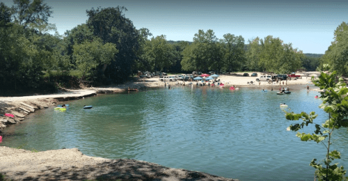 A sunny beach scene with people swimming and relaxing by a calm river, surrounded by trees and parked cars.