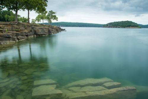 Serene lake view with rocky shoreline, trees, and a distant island under a cloudy sky.