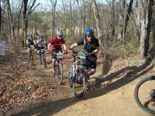 A group of mountain bikers navigating a dirt trail through a wooded area on a sunny day.