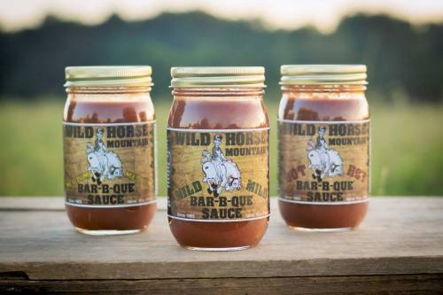 Three jars of Wild Horse Mountain Bar-B-Q Sauce on a wooden surface, set against a blurred green field background.