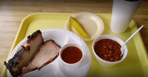 A tray with sliced smoked meat, barbecue sauce, baked beans, a pickle, and a drink in a cup.