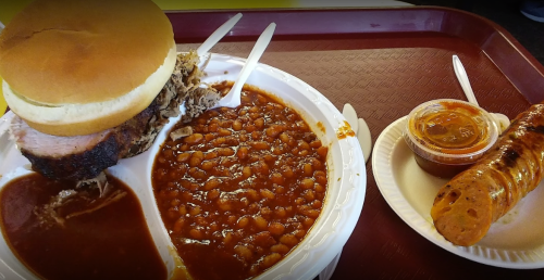 A plate with pulled pork sandwich, baked beans, and a sausage link, served on a tray.