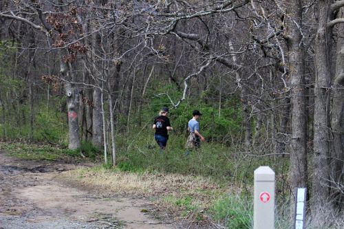 Two people walking through a wooded area, surrounded by trees and greenery, with a trail marker visible nearby.