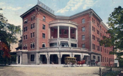 Historic brick hotel with a large porch, horse-drawn carriage, and trees in the foreground, set against a blue sky.