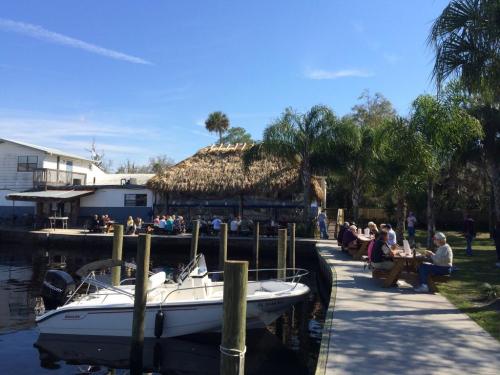 A waterfront scene with a thatched-roof restaurant, people dining outside, and a boat docked nearby under a clear blue sky.