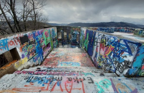 Colorful graffiti covers the walls and stairs of a weathered structure, with a cloudy landscape in the background.