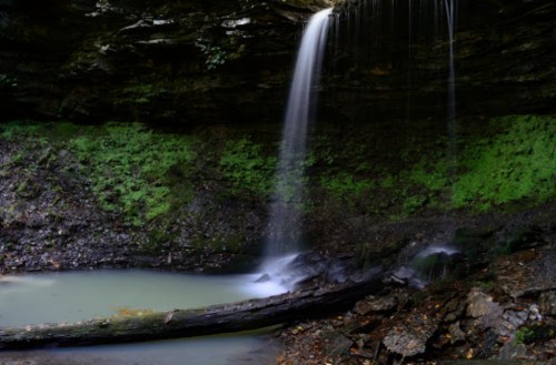 A serene waterfall cascades into a tranquil pool, surrounded by lush greenery and rocky terrain.