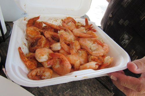 A takeout container filled with seasoned shrimp, held by a person, on a wooden table.
