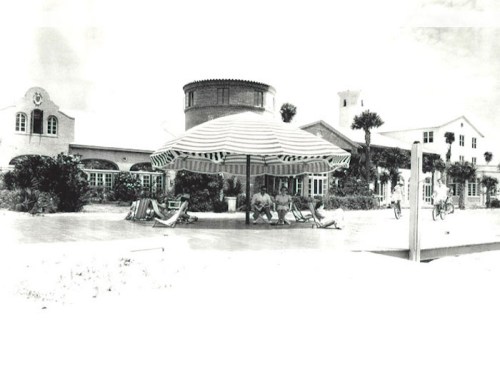 Black and white image of a beach scene with an umbrella, lounge chairs, and a coastal building in the background.