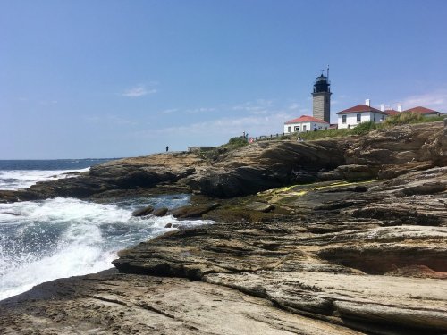 A rocky coastline with waves crashing, featuring a lighthouse and buildings under a clear blue sky.