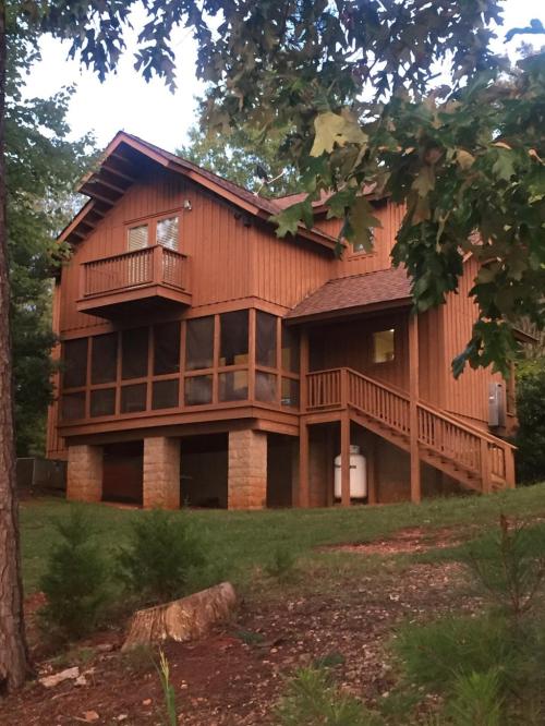 A wooden house with a screened porch, balcony, and stone foundation, surrounded by trees and greenery.