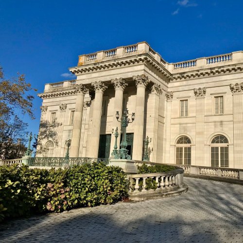 A grand, classical-style building with columns and sculptures, surrounded by greenery under a clear blue sky.