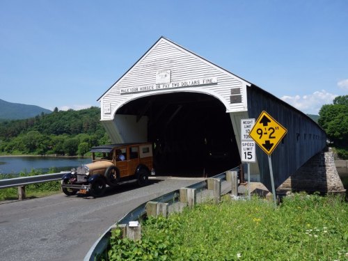 A vintage car drives through a covered bridge surrounded by greenery and a calm river under a clear blue sky.