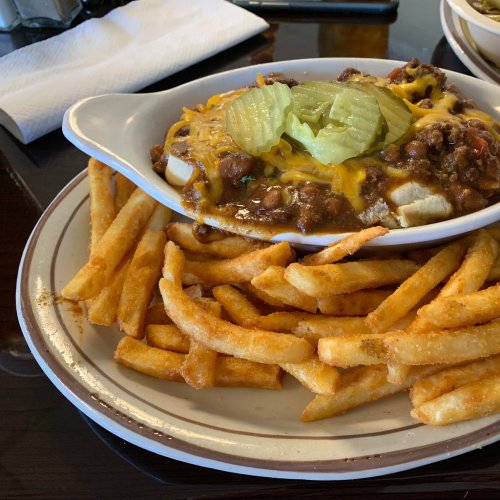 A plate of fries with a dish of chili topped with cheese and pickles.