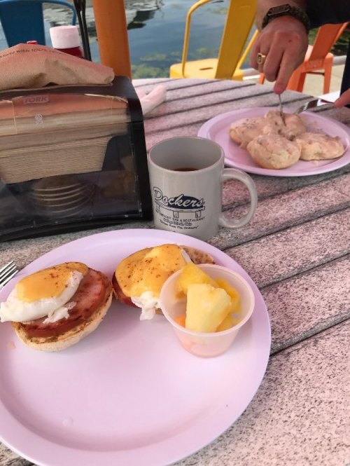 A breakfast spread featuring eggs Benedict, a fruit cup, and coffee on a table by the water.