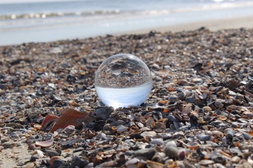 A glass sphere resting on a beach covered in seashells, reflecting the ocean and sky in the background.