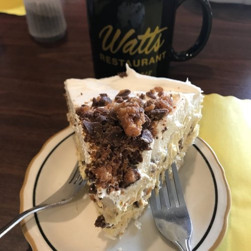 A slice of cake topped with nuts and frosting, served on a plate with two forks and a coffee mug in the background.