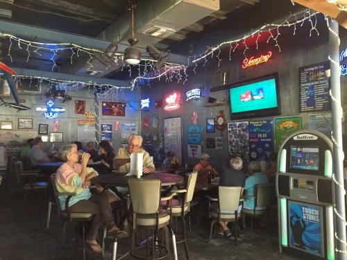 A lively bar interior with neon signs, patrons seated at tables, and a TV displaying content in the background.