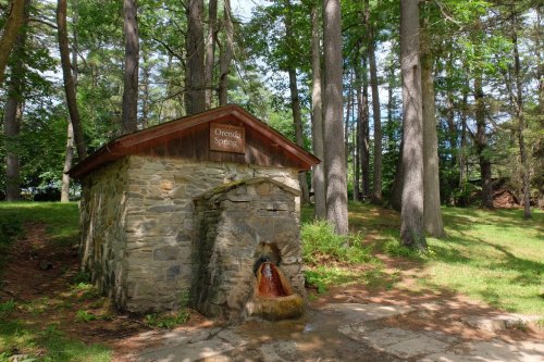 A stone structure labeled "Orenda Spring" surrounded by trees, with water flowing from its spout onto the ground.