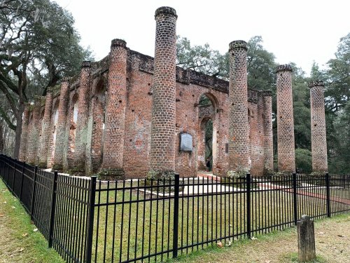 Ruins of a brick structure with tall columns, surrounded by a black fence and greenery, under a cloudy sky.