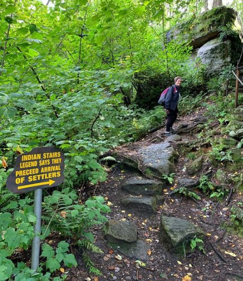 A person walks up a rocky path surrounded by lush greenery, with a sign reading "Indian Stairs" nearby.