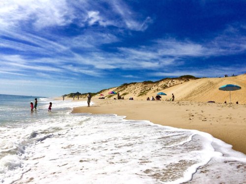 A sunny beach scene with people enjoying the water, colorful umbrellas, and sandy dunes under a blue sky.