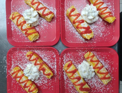Four red plates with rolled desserts topped with whipped cream and drizzled with red sauce, sprinkled with powdered sugar.
