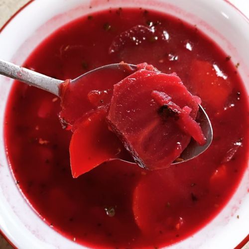 A spoonful of vibrant red beet soup with chunks of beets and vegetables in a white bowl.