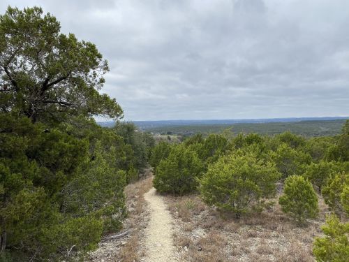 A winding dirt path leads through green shrubs and trees, with a cloudy sky and distant hills in the background.