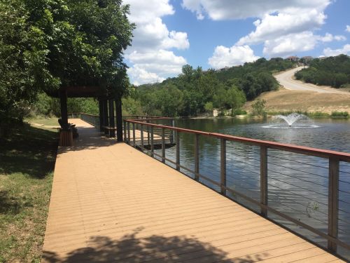 A wooden walkway beside a calm lake, with trees and a fountain in the background under a partly cloudy sky.